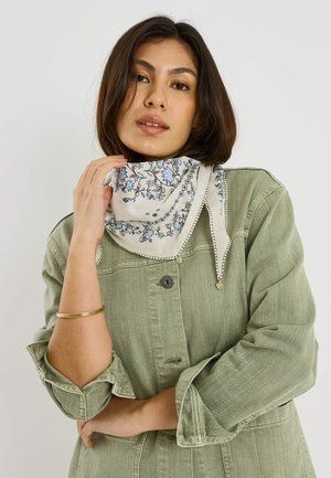 Femme portant une veste verte à boutons et une écharpe crème à motifs floraux bleus, touchant l'écharpe près du cou, sur un fond uni.