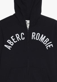 Mørk marineblå zip-up hoodie lavet af blødt stof med hvid broderet tekst "ABERCROMBIE" i et buet design. Indeholder hætte og frontlommer.