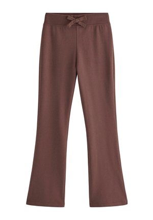 Smilende pige med langt blond hår iført en hvid blomstret kortærmet top, brune bukser og hvide sneakers, stående på en ensfarvet baggrund.