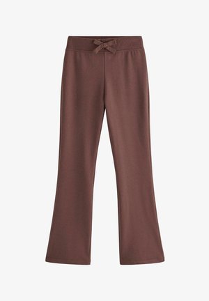 Smilende pige med langt blond hår iført en hvid blomstret kortærmet top, brune bukser og hvide sneakers, stående på en ensfarvet baggrund.