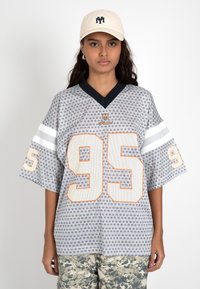 Joven mujer con gorra beige, camiseta deportiva gris con patrón y el número 95, y pantalones de camuflaje, de pie frente a un fondo blanco.