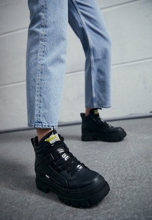Czarne, wysokie sneakersy z czarnej skóry z masywną podeszwą, sznurowane, z żółtą metką oraz teksturowanymi akcentami. Nosi się je z jasnoniebieskimi dżinsami.
