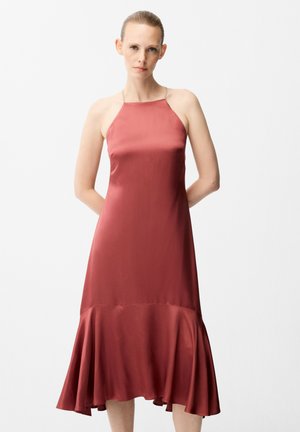 Femme à la peau claire et aux cheveux attachés, portant une robe midi sans manches couleur rouille avec un ourlet évasé, debout, les mains dans le dos.