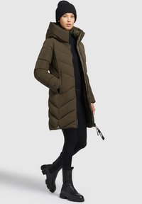 Olijfgroene pufferjas met diagonale gestikte patronen, capuchon en ritssluiting. Gecombineerd met zwarte legging en zwarte laarzen tot de knie.