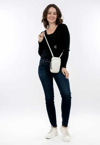 Bílá crossbody taška s hladkým povrchem, zapínáním na zip a detailem střapce, v kombinaci s černým svetrem a tmavými džíny.