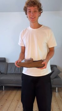 Braune Wildlederslipper, die von einer Person gehalten werden, mit einer runden Zehenpartie, minimalen Nähten und einem leicht erhöhten dekorativen Riemen.