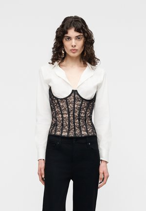 Γυναίκα με κατσαρά μαλλιά φοράει λευκό πουκάμισο με γιακά κάτω από μαύρο κορσέ από δαντέλα και μαύρο παντελόνι, στέκεται μπροστά σε ανοιχτό γκρι φόντο.