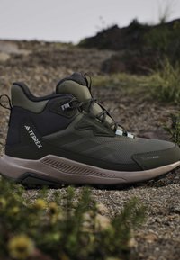 Dark green and black Terrex hiking boot with sturdy sole on rocky outdoor terrain with sparse vegetation.
