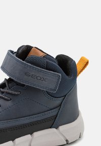 Zapatilla alta de cuero azul y malla con tira de Velcro, suela de goma gris y lengüeta de acento naranja en el talón. Presenta costuras en contraste.