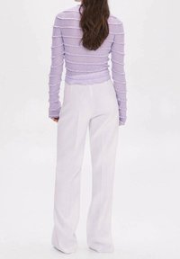 Femme aux longs cheveux foncés portant un haut à manches longues texturé couleur lavande et un pantalon blanc taille haute à jambes larges, debout de dos.