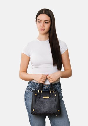 Junge Frau mit langen dunklen Haaren, die ein weißes T-Shirt und Jeans trägt und eine schwarze Handtasche mit goldenen Akzenten vor einem schlichten Hintergrund hält.
