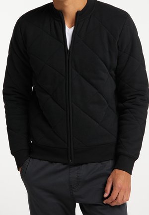 Homme portant une veste matelassée noire zippée par-dessus une chemise blanche avec un pantalon foncé, mains dans les poches, debout devant un fond uni.