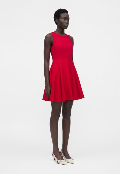 Γυναίκα που στέκεται φορώντας ένα αμάνικο κόκκινο φόρεμα με φουρό, σε συνδυασμό με λευκές μυτερές γόβες slingback σε απλό ανοιχτό γκρι φόντο.