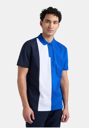 Giovane uomo che indossa una polo a maniche corte con blocchi di colore verticali in blu navy, bianco e blu reale, in piedi davanti a uno sfondo uniforme.