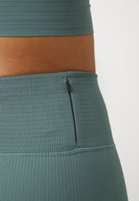 Curea sport verde albastru dungi, cu un buzunar cu fermoar pe lateral, oferind un accent de design funcțional. Textură netedă cu o formă ajustată.