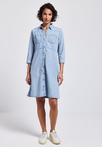Robe chemise en denim bleu clair avec des manches longues, fermeture à boutons, deux poches poitrine et une jupe évasée, assortie à des baskets blanches.