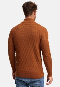 Homme portant un pull en maille texturée couleur rouille et un jean bleu, vu de dos sur un fond uni.