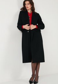 Palton lung negru din amestec de lână, cu un design măsurat, având un guler cu tăietură, nasturi frontali și buzunare mari. Textură netedă.