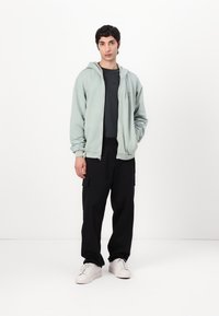 Felpa verde chiaro con zipper sopra una maglietta grigia scura, abbinata a pantaloni cargo neri e sneakers bianche. Tessuto morbido, vestibilità rilassata.