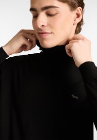 Schwarzer Rollkragenpullover in ribbed Struktur, mit einem dezenten Hundelogo auf der linken Brust und umgeschlagenem Stoff am Kragen.