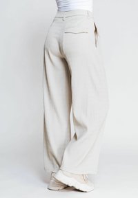 Beige weit geschnittene Hose aus strukturiertem Stoff. Verfügt über eine Gesäßtasche und dezente vertikale Streifen, kombiniert mit hellen Sneakers.