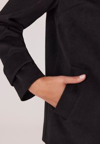 Czarna kurtka o teksturowanym wykończeniu. Zbliżenie na rękaw i boczną kieszeń, ukazujące dopasowany design i czyste linie.