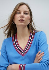 Blå tröja med djup V-ringning, med röd och marinblå randig kant; broderat hjärta på bröstet; slät stickad textur.