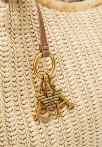 Beige vävd stråväska med en guldfärgad metallberlock med logotyp, fäst vid en brun läderrem och en cirkulär metallring.