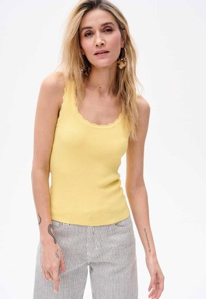 Żółty podkoszulek z prążkowaniem i falbaniastym dekoltem, zestawiony z szerokimi spodniami w paski. Widoczne tatuaże na jednym ramieniu.