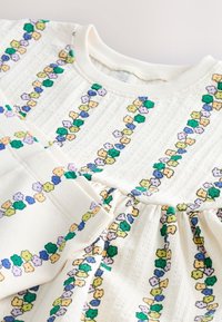 Vestito bianco in cotone con tessuto strutturato, decorato con ricami floreali gialli, viola, verdi e blu, disposti a strisce verticali.