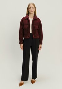 Bordeaux cropped wollen jack met twee opgestikte zakken, knoopsluiting, gedragen over een lichte top, gecombineerd met zwarte broeken en bruine hakken.