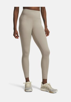 Vrouw draagt beige high-waist legging met zijzakken en crème sneakers, staand op een witte achtergrond.