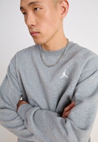 Felpa grigia con maniche lunghe, caratterizzata da un piccolo logo Jumpman bianco sul petto. Il tessuto sembra morbido con un taglio casual. Collana in argento visibile.