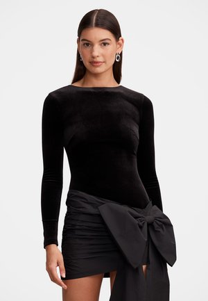 Jeune femme aux longs cheveux raides portant une robe en velours noir ajustée avec des manches longues et un grand nœud à la taille.