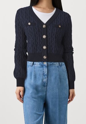 Cardigan court en maille torsadée bleu marine avec des boutons dorés, porté sur un top blanc, associé à un jean bleu clair ample et taille haute.