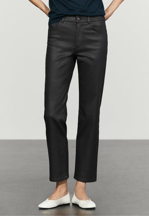 Person trägt schwarze Slim-Fit-Hose und weiße Ballerinas, steht mit den Händen hinter dem Rücken auf einem grauen Boden.