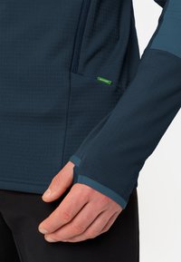 Maglia a maniche lunghe sintetica, di colore blu navy e con texture, dotata di tasca con zip e aperture per i pollici; presenta un'etichetta verde con logo sul lato.