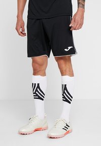 Mustat urheilushortsit, joissa on valkoiset yksityiskohdat, yhdistettynä polven ylle ulottuviin valkoisiin sukkiin, joissa on mustia viistoja raitoja sekä valkoisiin urheilukenkään.