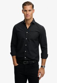 Zwarte lange mouwen shirt met knoop-kraag, witte knopen en een subtiele logo. G gedragen met zwarte broek en een gevlochten zwarte riem.