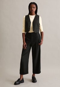 Schwarze maßgeschneiderte weit geschnittene Hose, eine olivgrüne taillierte Weste mit Knöpfen und ein hellgelbes Langarmoberteil. Schwarze Slipper runden das Outfit ab.