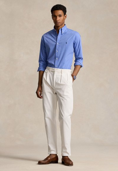 Bărbat purtând o cămașă albastră cu nasturi, pantaloni albi plisati, mocasini maro din piele și un ceas de mână, stând în fața unui fundal neutru.