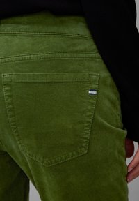 Pantaloni in corduroy verde con una consistenza morbida, dotati di tasche posteriori e un piccolo etichetta a righe vicino alla cucitura della tasca.