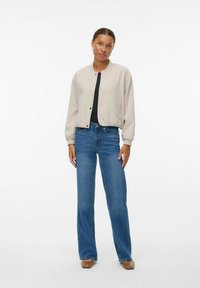 Vrouw die staat in een beige bomberjack, zwart topje, blauwe wide-leg jeans en bruine vierkante schoenen op een effen witte achtergrond.