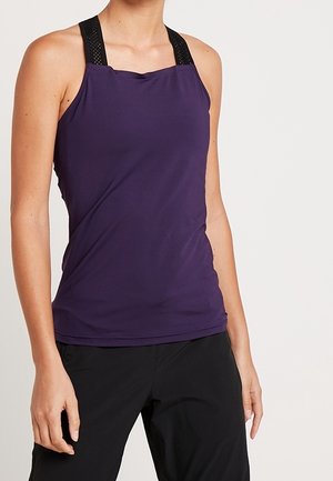 Kobieta ubrana w bezrękawnik w kolorze fioletowym z czarnymi siateczkowymi paskami oraz czarne spodnie, widoczna od szyi do bioder na jednolitym tle.