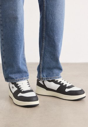 Personne portant un jean bleu et des baskets noir et blanc, debout sur un sol gris clair sur un fond blanc uni.