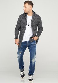 Knit gray cardigan with a shawl collar over a white t-shirt, paired with distressed blue jeans and black sneakers, standing on a neutral background.