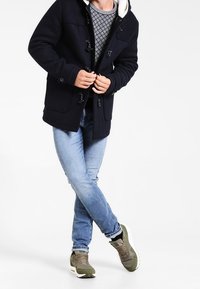 Persona in cappotto blu con bottoni a toggle, maglione grigio a fantasia, jeans blu chiaro e sneakers verdi, in piedi con le gambe incrociate.