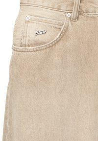 Beige farkkuhousut, joissa on hieman teksturoitu pinta, etutaskussa pieni hopeinen logo ja klassiset ompeluyksityiskohdat.