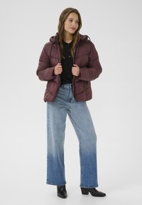 Mørk burgunder pufferjakke med hætte, der har et quiltet design og sorte snore. Parret med højtaljede lyseblå jeans og sorte støvler.