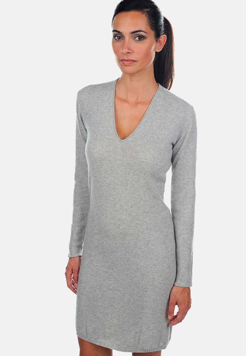 Femme aux cheveux bruns en queue de cheval, portant une robe en tricot gris clair à manches longues et décolleté en V, se tenant devant un fond blanc uni.